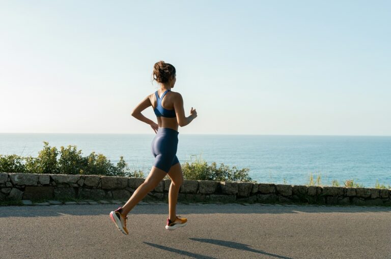 Uma mulher correndo ao longo de um caminho ao ar livre, com o mar calmo ao fundo e um céu claro. Ela está vestindo um top esportivo azul, shorts curtos combinando e tênis coloridos, enquanto mantém uma postura ativa e dinâmica.